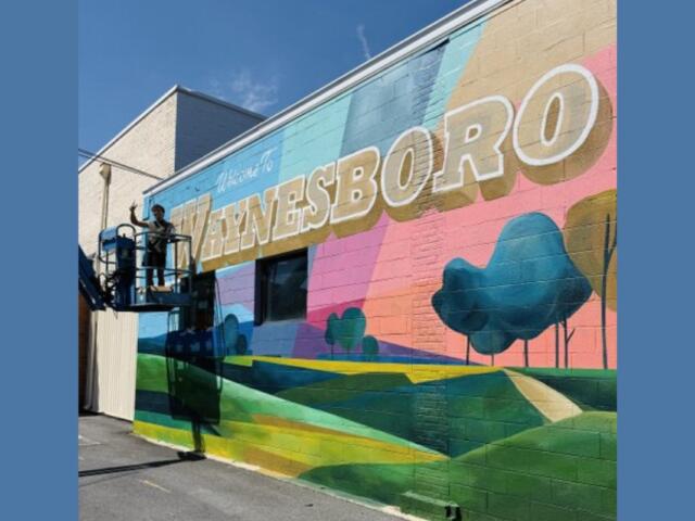 Worker on a lift painting a colorful mural on a brick wall that reads 'WAYNESBORO'.