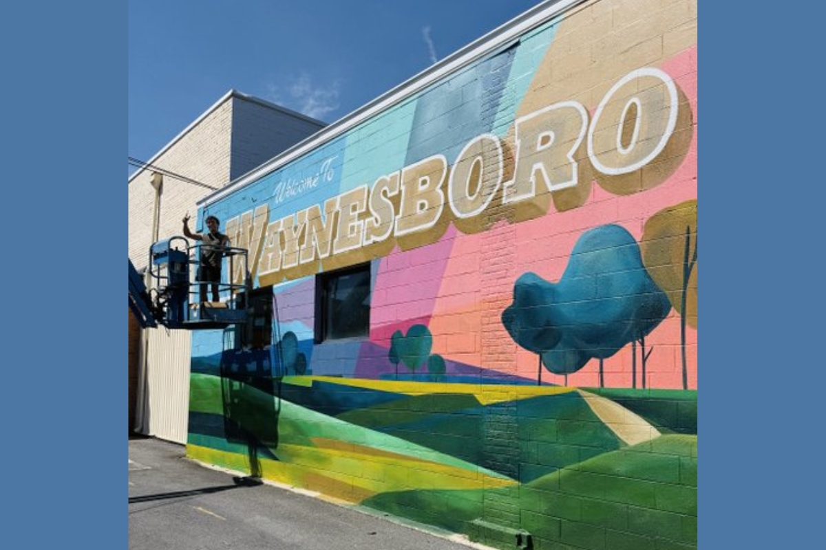 Worker on a lift painting a colorful mural on a brick wall that reads 'WAYNESBORO'.