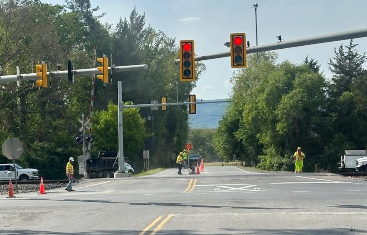 Railroad crossing with red signal lights, orange cones, and workers in high‑visibility vests directing traffic on a sunny day.