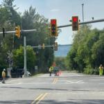 Railroad crossing with red signal lights, orange cones, and workers in high‑visibility vests directing traffic on a sunny day.