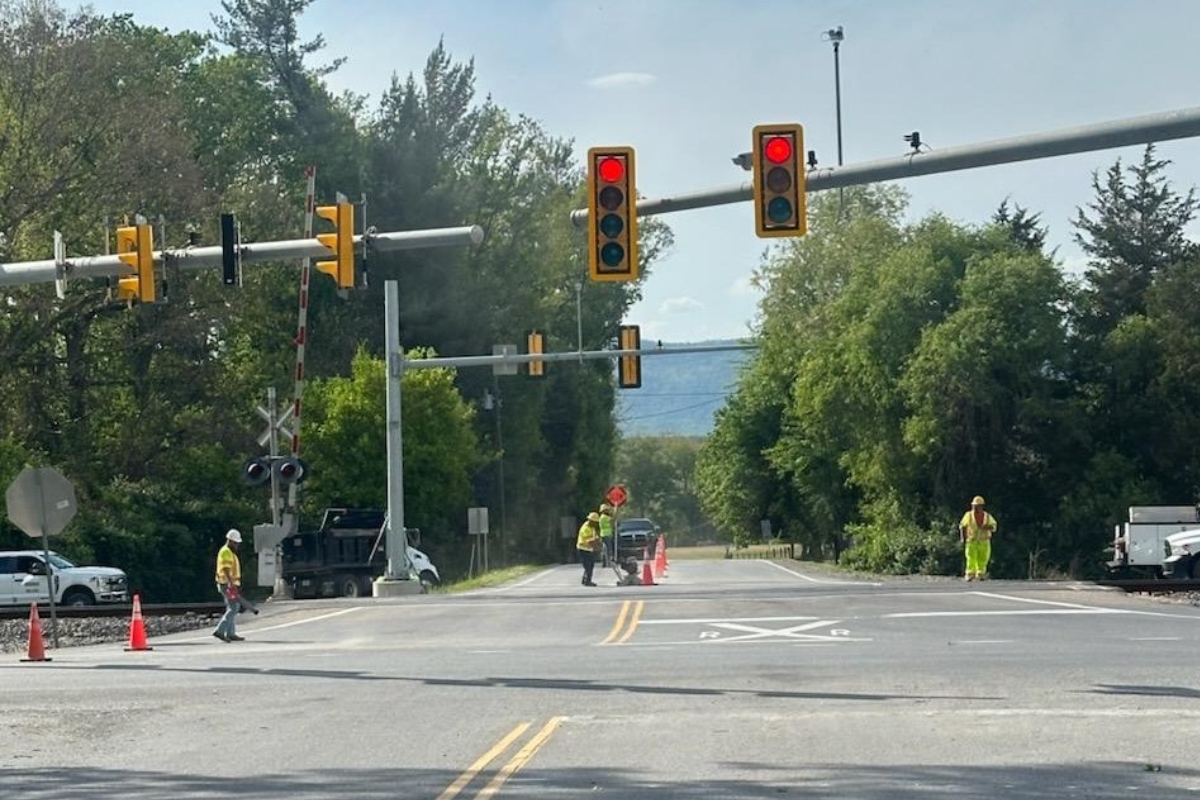 Railroad crossing with red signal lights, orange cones, and workers in high‑visibility vests directing traffic on a sunny day.