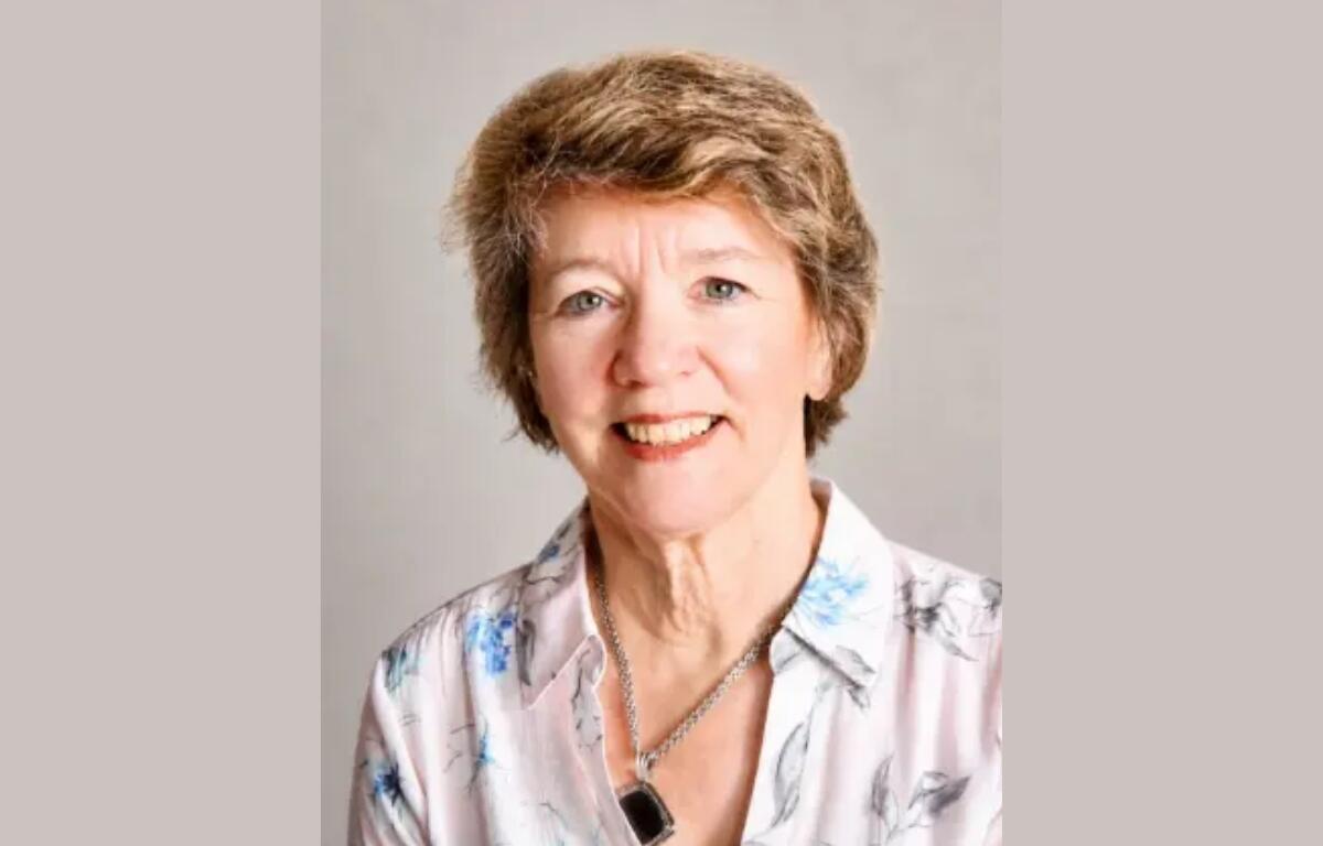 Portrait of a smiling older woman with short light brown hair, wearing a floral blouse and necklace.