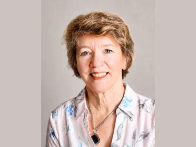 Portrait of a smiling older woman with short light brown hair, wearing a floral blouse and necklace.