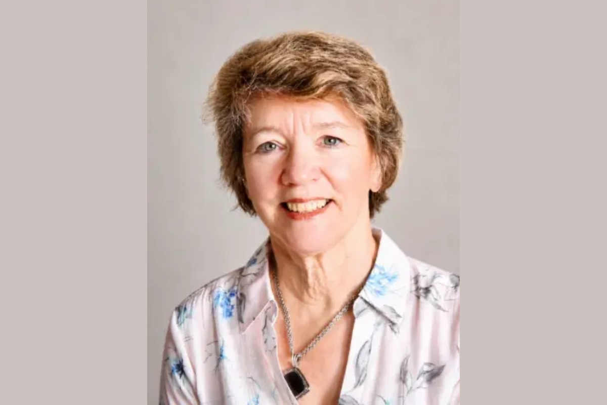 Portrait of a smiling older woman with short light brown hair, wearing a floral blouse and necklace.
