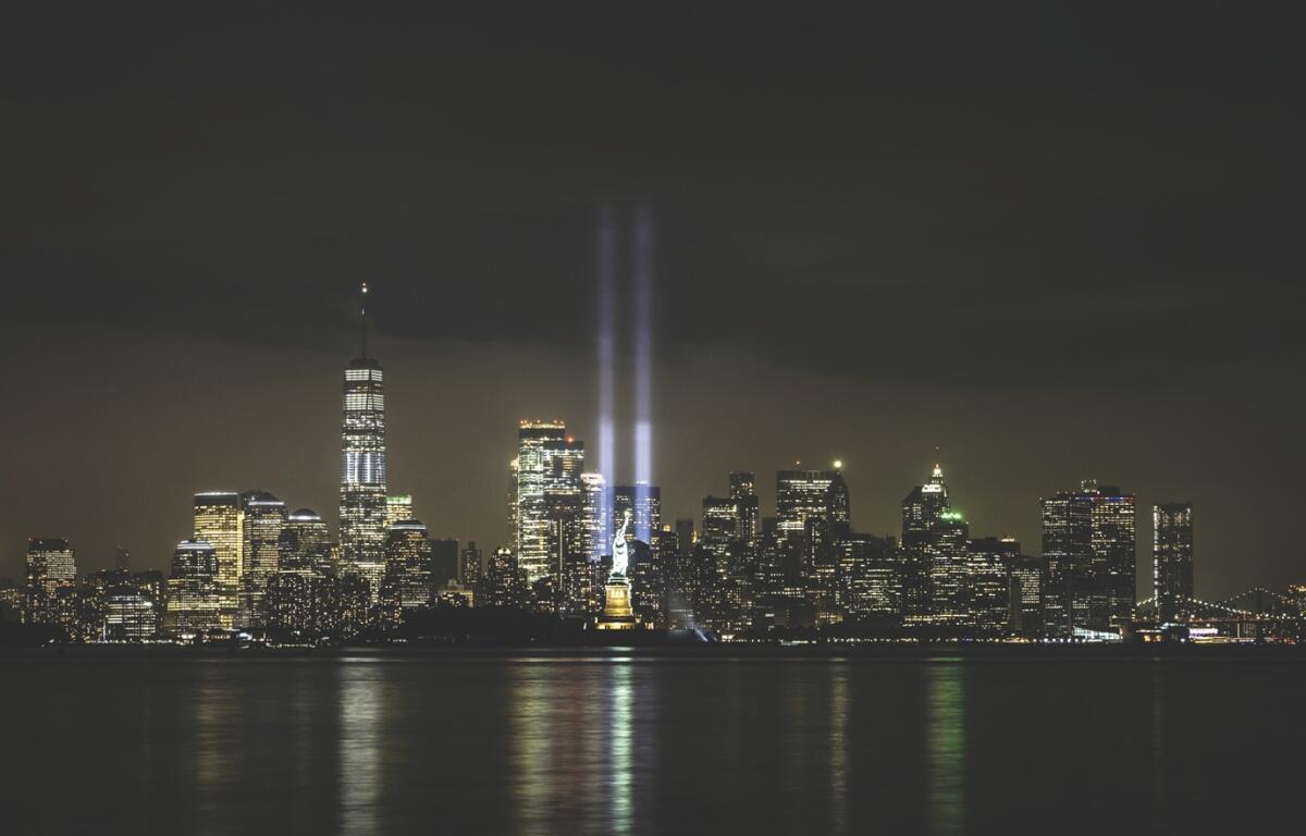 Nighttime city skyline of Manhattan with lit buildings and two blue memorial beams rising from the World Trade Center site.