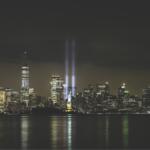 Nighttime city skyline of Manhattan with lit buildings and two blue memorial beams rising from the World Trade Center site.