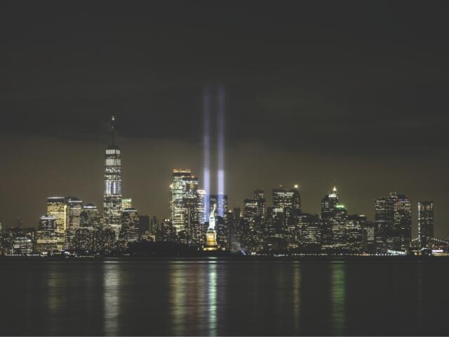 Nighttime city skyline of Manhattan with lit buildings and two blue memorial beams rising from the World Trade Center site.