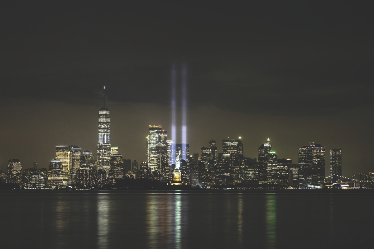 Nighttime city skyline of Manhattan with lit buildings and two blue memorial beams rising from the World Trade Center site.