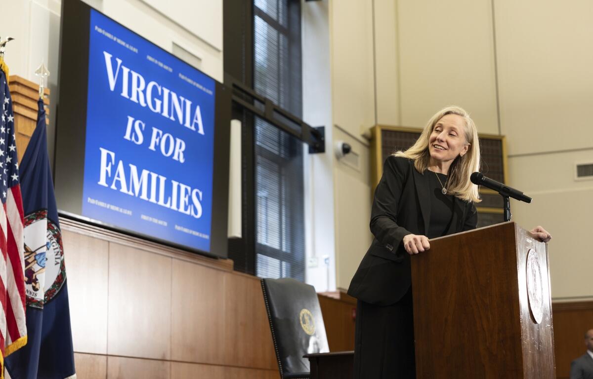 Woman in a black suit stands at a wooden podium delivering a speech, with a blue poster reading 'Virginia Is For Families' in the background and flags nearby.
