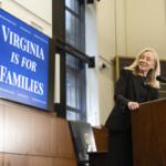 Woman in a black suit stands at a wooden podium delivering a speech, with a blue poster reading 'Virginia Is For Families' in the background and flags nearby.