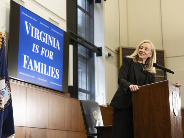 Woman in a black suit stands at a wooden podium delivering a speech, with a blue poster reading 'Virginia Is For Families' in the background and flags nearby.