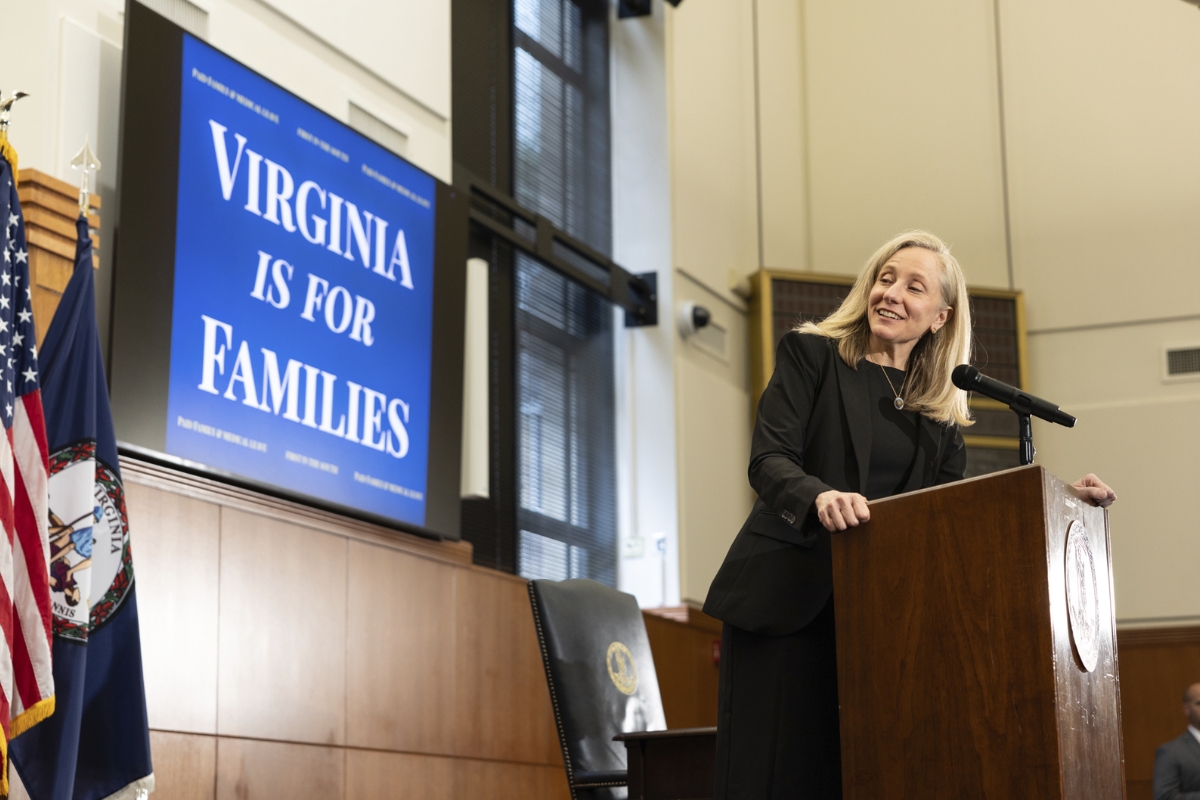 Woman in a black suit stands at a wooden podium delivering a speech, with a blue poster reading 'Virginia Is For Families' in the background and flags nearby.