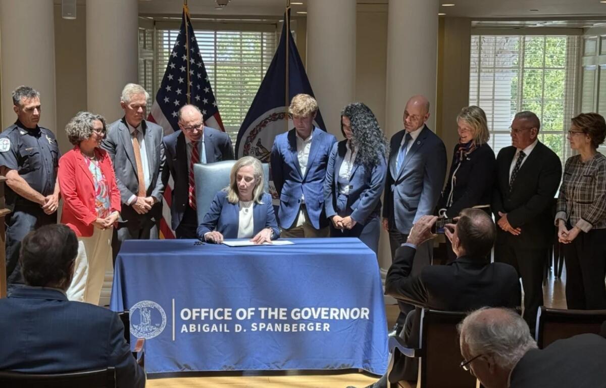 Governor signs document at a desk covered with a blue cloth reading 'Office of the Governor' as officials stand behind him/her with flags in the background.