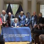 Governor signs document at a desk covered with a blue cloth reading 'Office of the Governor' as officials stand behind him/her with flags in the background.