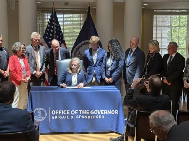 Governor signs document at a desk covered with a blue cloth reading 'Office of the Governor' as officials stand behind him/her with flags in the background.