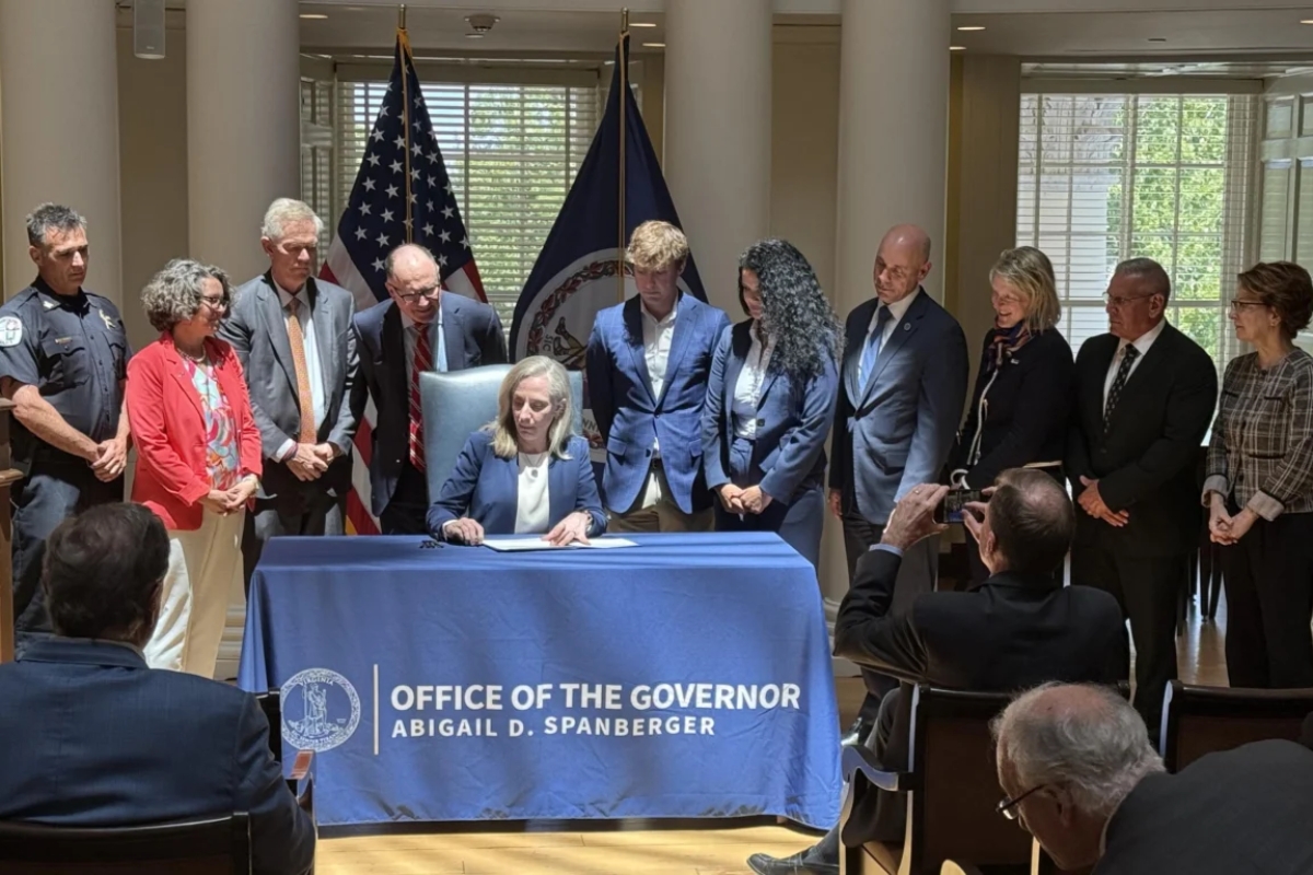 Governor signs document at a desk covered with a blue cloth reading 'Office of the Governor' as officials stand behind him/her with flags in the background.
