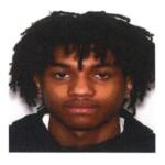 Head-and-shoulders portrait of a young man with dark dreadlocks, wearing a black top, neutral expression against a white background.