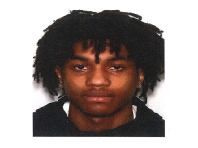 Head-and-shoulders portrait of a young man with dark dreadlocks, wearing a black top, neutral expression against a white background.