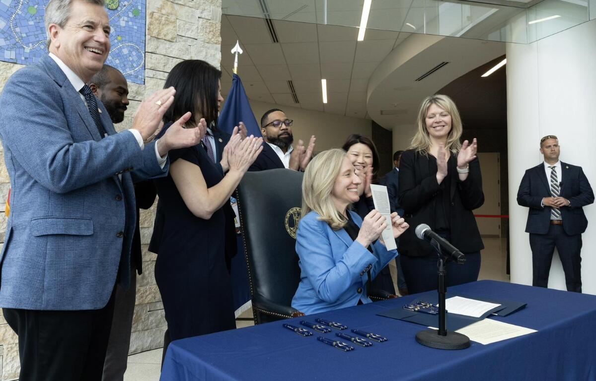 Woman in a blue blazer signs a document at a table as colleagues applaud in a bright lobby ceremony.