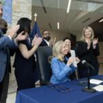 Woman in a blue blazer signs a document at a table as colleagues applaud in a bright lobby ceremony.