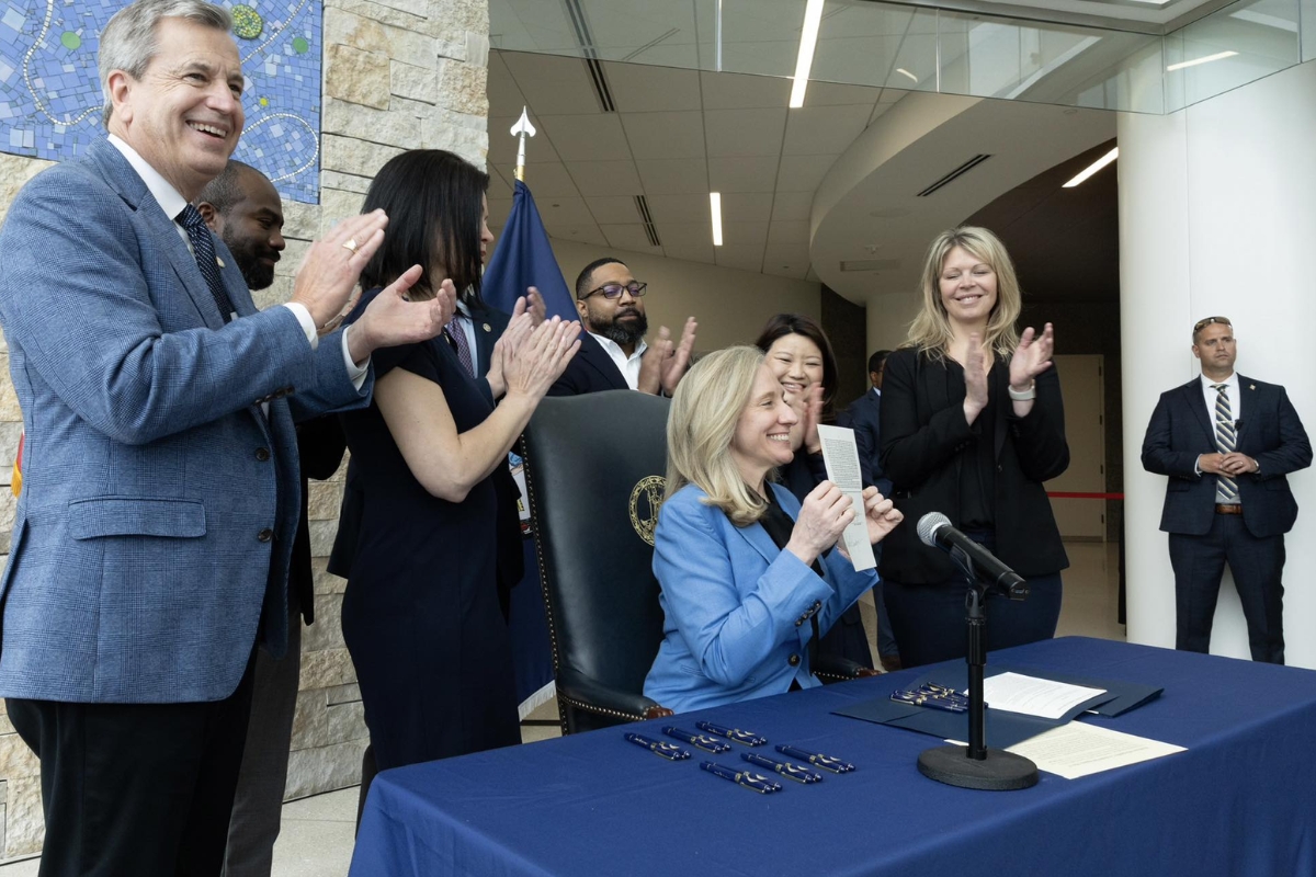 Woman in a blue blazer signs a document at a table as colleagues applaud in a bright lobby ceremony.