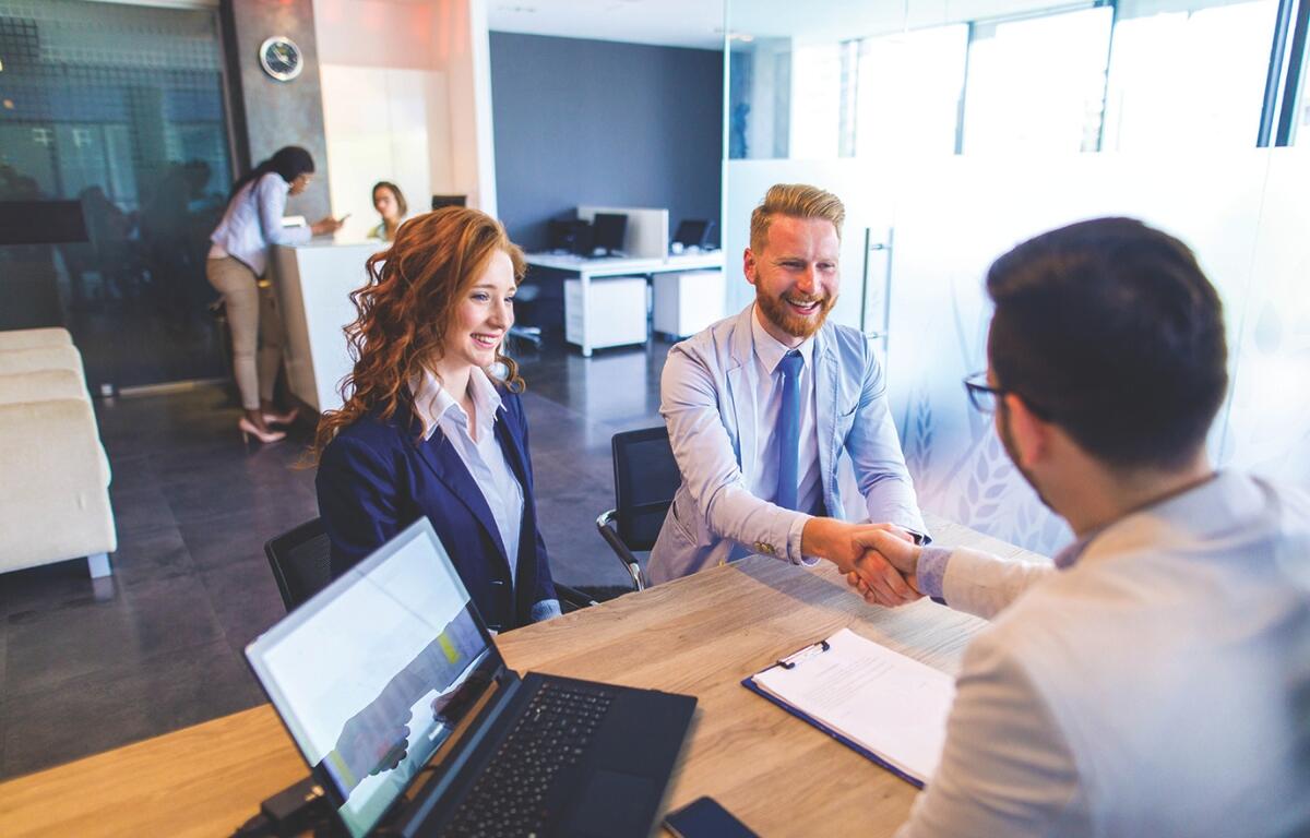 Two business professionals shake hands across a wooden desk in a bright, modern office, with a laptop and clipboard nearby.