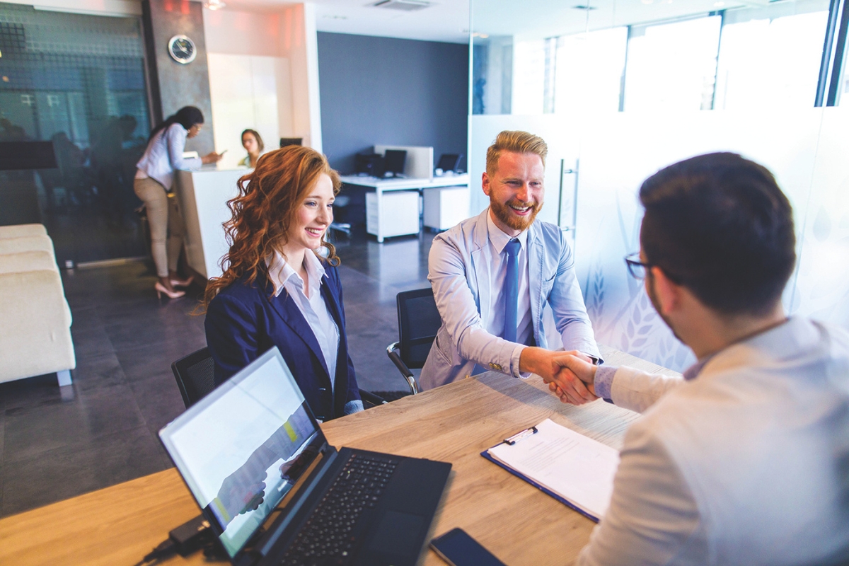 Two business professionals shake hands across a wooden desk in a bright, modern office, with a laptop and clipboard nearby.