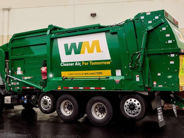 Green WM refuse truck with 'Cleaner Air, For Tomorrow' branding parked beside a building, showing large wheels and rear loading area.