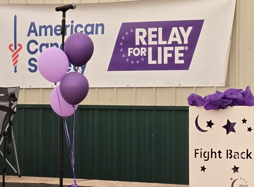 Microphone stand with purple balloons in front of a Relay For Life and American Cancer Society banner at a fundraiser event.