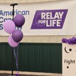 Microphone stand with purple balloons in front of a Relay For Life and American Cancer Society banner at a fundraiser event.