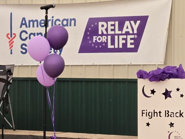 Microphone stand with purple balloons in front of a Relay For Life and American Cancer Society banner at a fundraiser event.