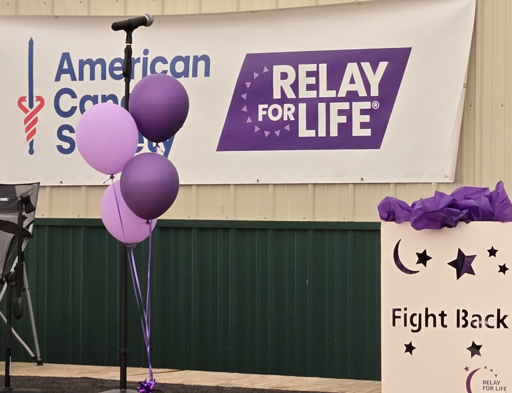Microphone stand with purple balloons in front of a Relay For Life and American Cancer Society banner at a fundraiser event.