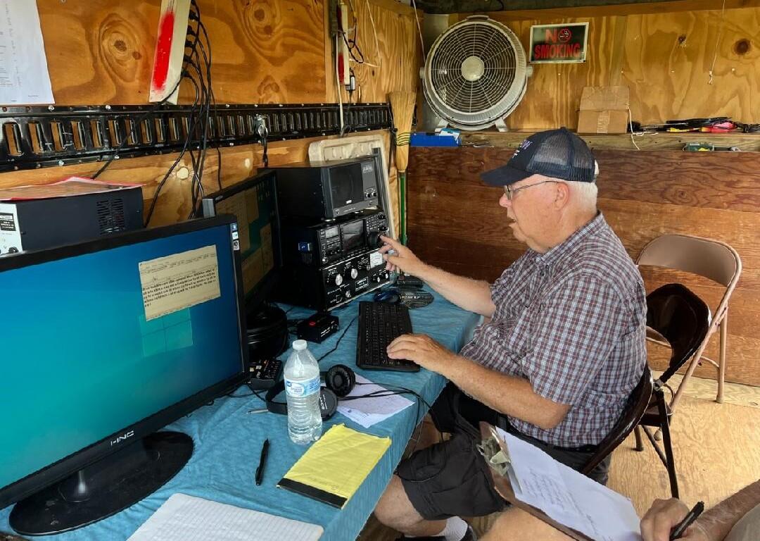 Older man wearing a cap sits at a workbench using vintage radio gear and a computer in a wooden shack.