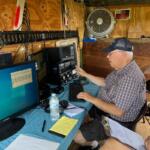 Older man wearing a cap sits at a workbench using vintage radio gear and a computer in a wooden shack.