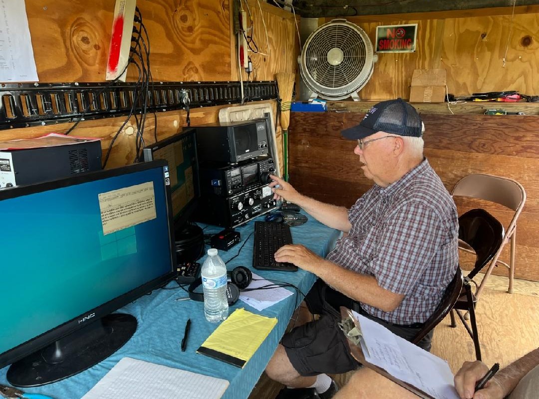 Older man wearing a cap sits at a workbench using vintage radio gear and a computer in a wooden shack.