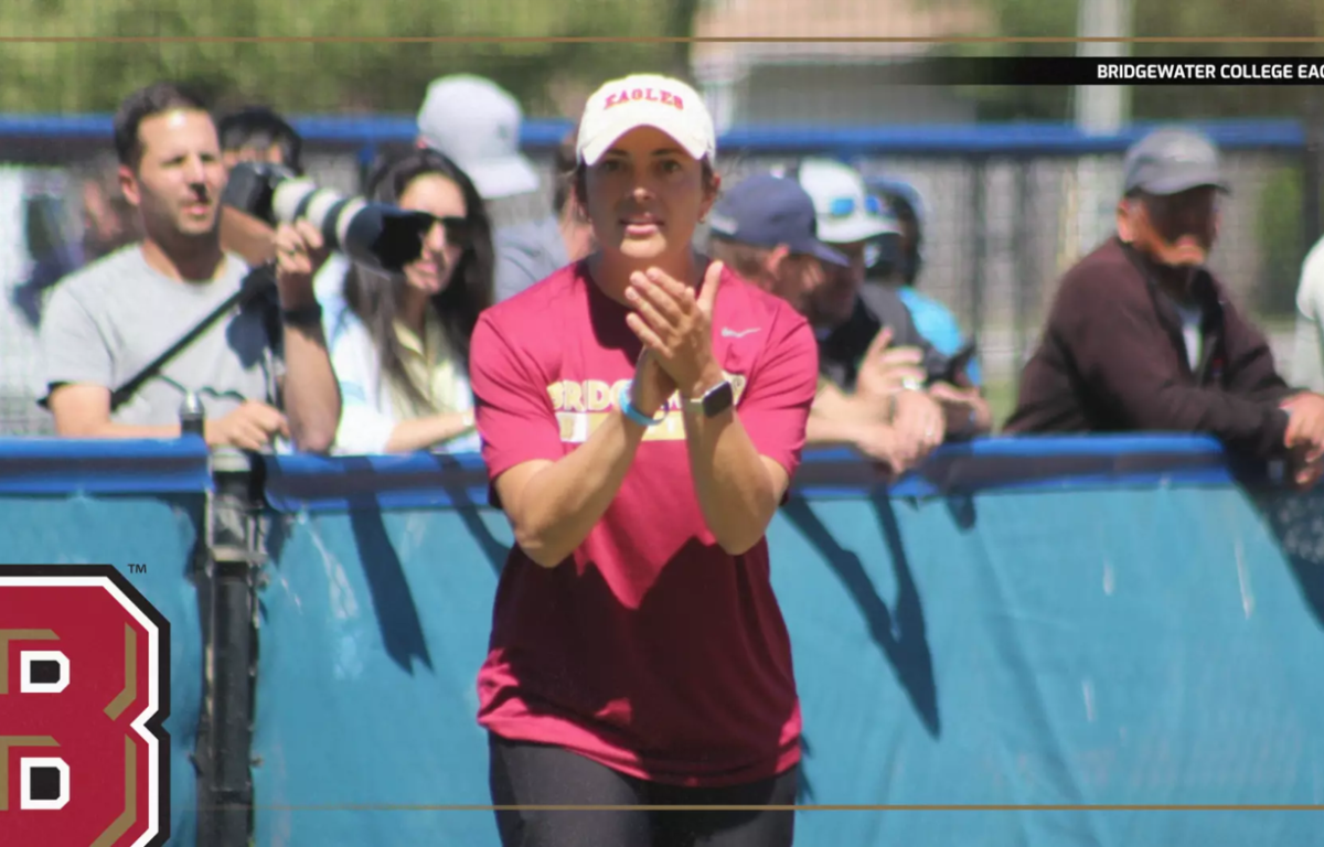 Female athlete in a maroon shirt and white cap claps at a blue barrier while spectators watch from behind.