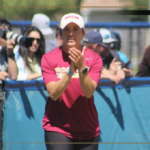Female athlete in a maroon shirt and white cap claps at a blue barrier while spectators watch from behind.