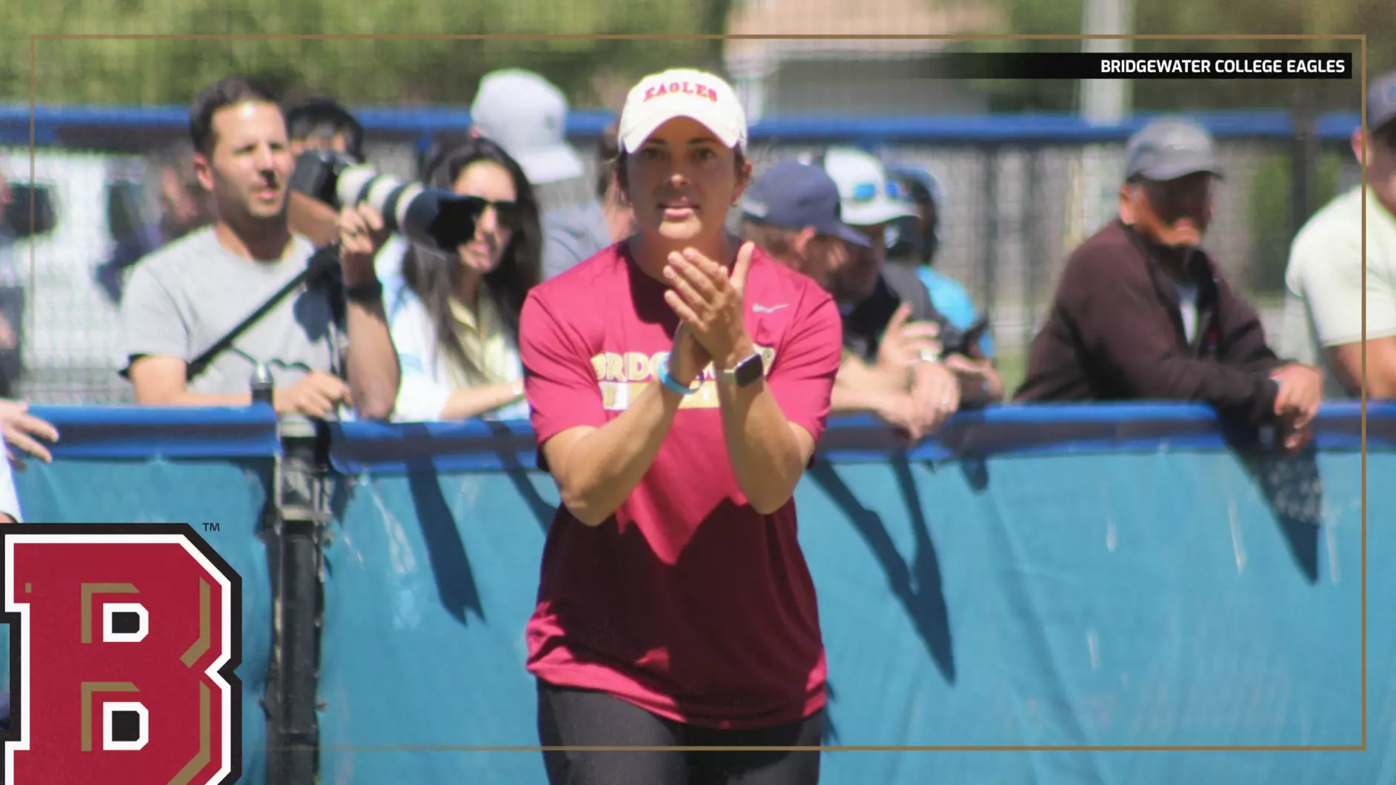 Female athlete in a maroon shirt and white cap claps at a blue barrier while spectators watch from behind.