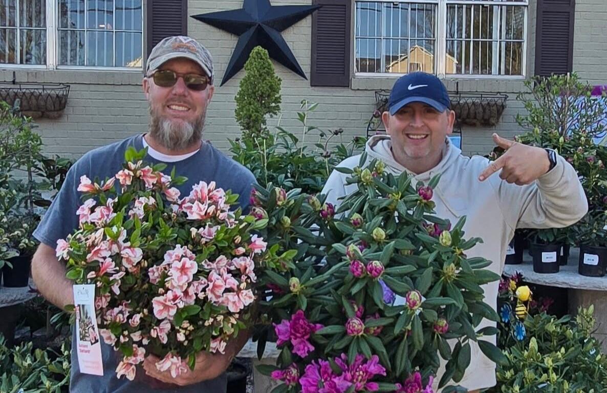 Two men stand outdoors in front of a house, each holding a flowering azalea shrub with pink and white blooms.