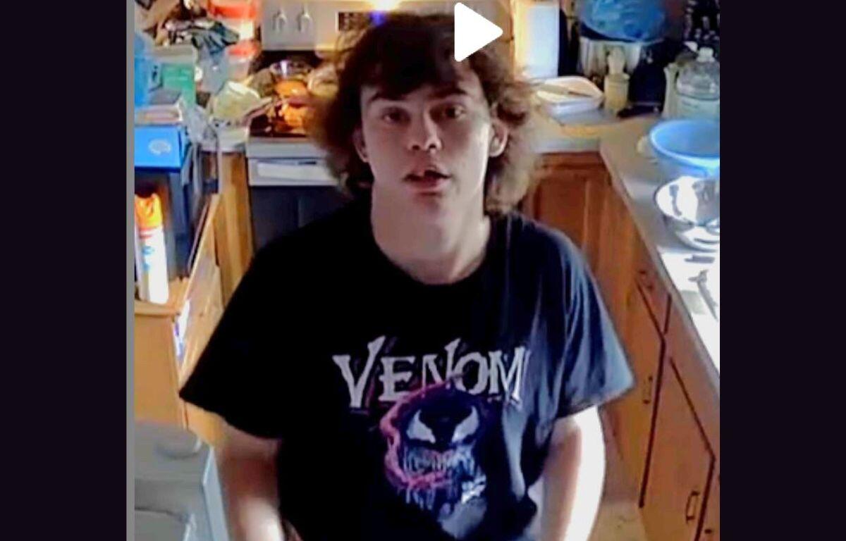 Young man with shoulder-length brown hair stands in a cluttered kitchen, wearing a black Venom t-shirt and looking at the camera.