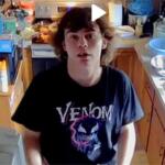 Young man with shoulder-length brown hair stands in a cluttered kitchen, wearing a black Venom t-shirt and looking at the camera.
