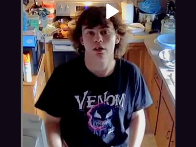 Young man with shoulder-length brown hair stands in a cluttered kitchen, wearing a black Venom t-shirt and looking at the camera.