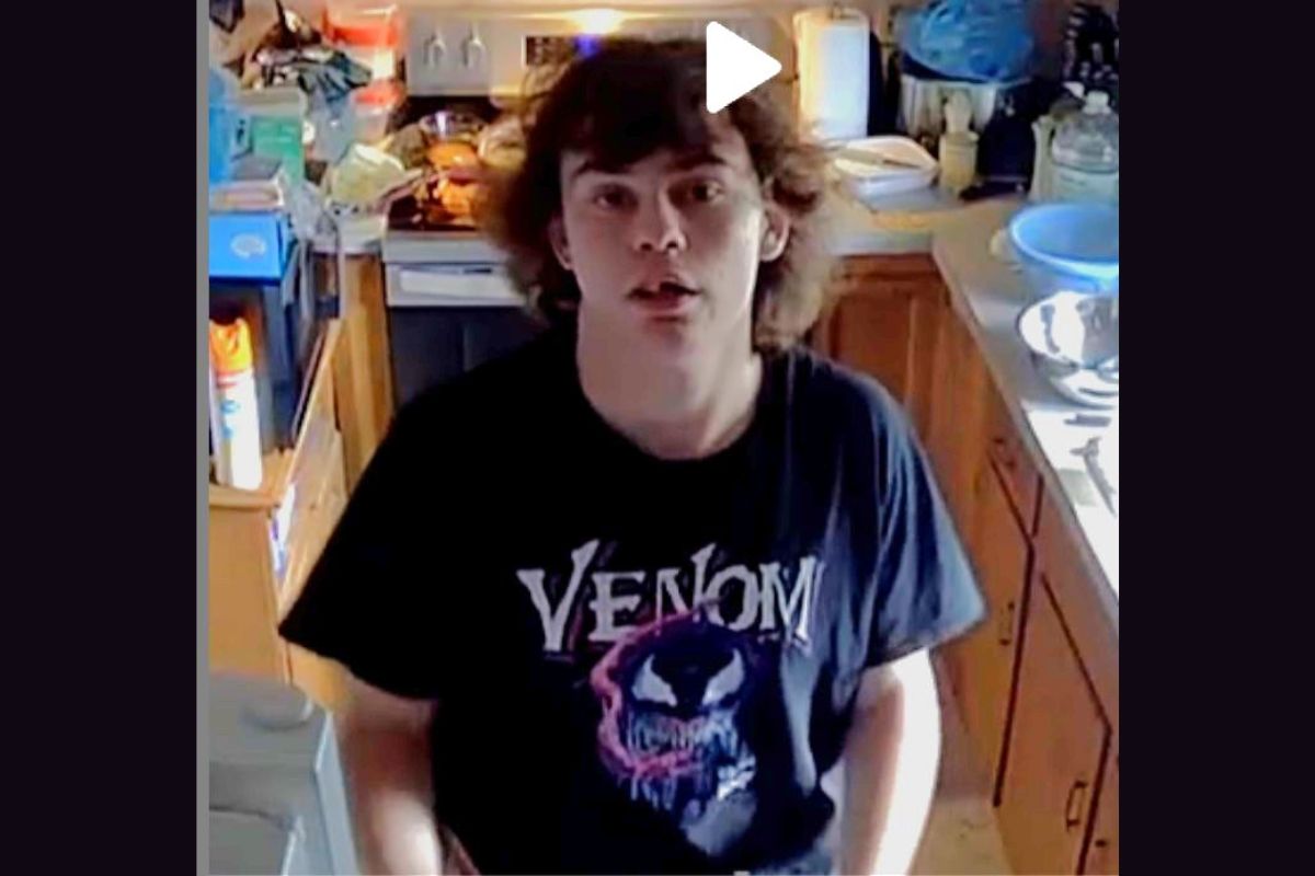 Young man with shoulder-length brown hair stands in a cluttered kitchen, wearing a black Venom t-shirt and looking at the camera.
