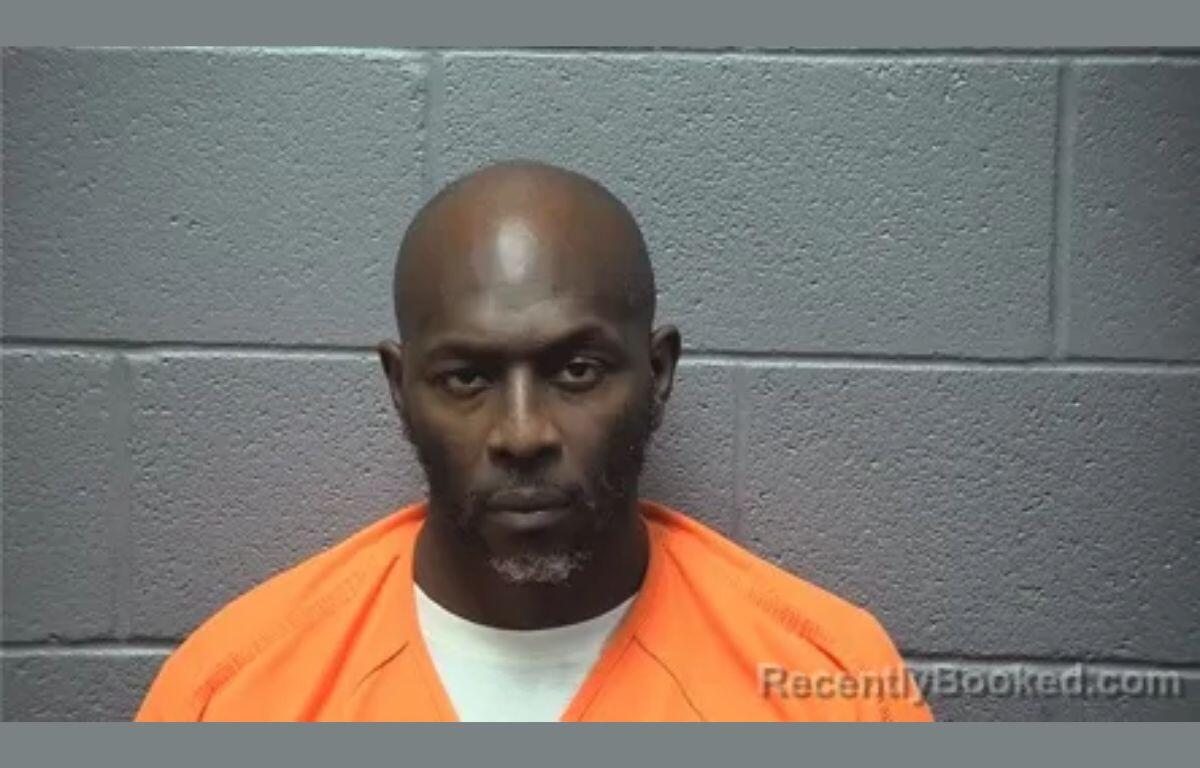Mugshot of a man in an orange jail jumpsuit against a gray concrete block wall.