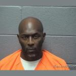 Mugshot of a man in an orange jail jump­suit against a gray concrete block wall.