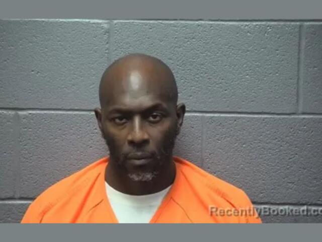 Mugshot of a man in an orange jail jump­suit against a gray concrete block wall.