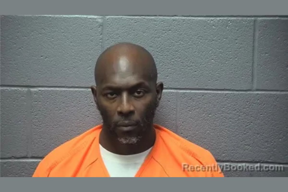 Mugshot of a man in an orange jail jump­suit against a gray concrete block wall.
