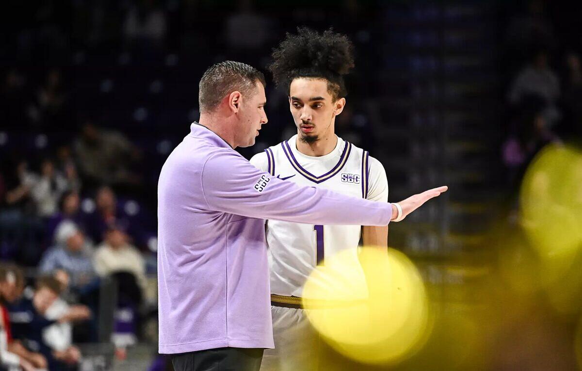 Basketball coach in a lavender jacket gestures to a player in a white jersey with number 1 during a timeout on the court, stands blurred in the background.