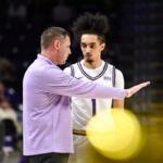 Basketball coach in a lavender jacket gestures to a player in a white jersey with number 1 during a timeout on the court, stands blurred in the background.