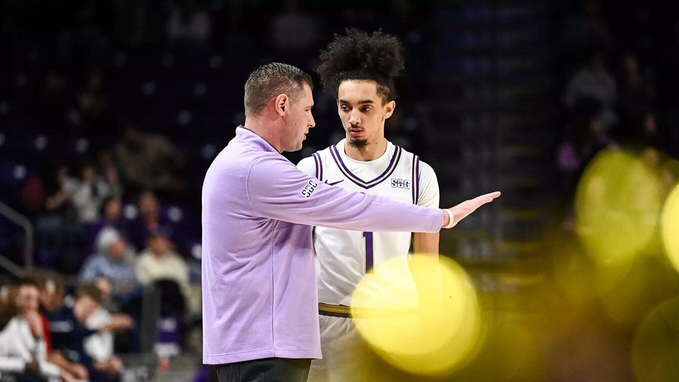 Basketball coach in a lavender jacket gestures to a player in a white jersey with number 1 during a timeout on the court, stands blurred in the background.